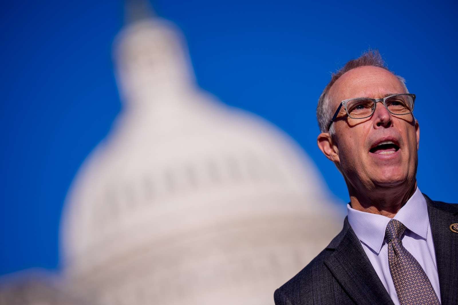 A white man with a receding hairline and glasses stands against a dark blue sky with the US Capitol building blurry in the background