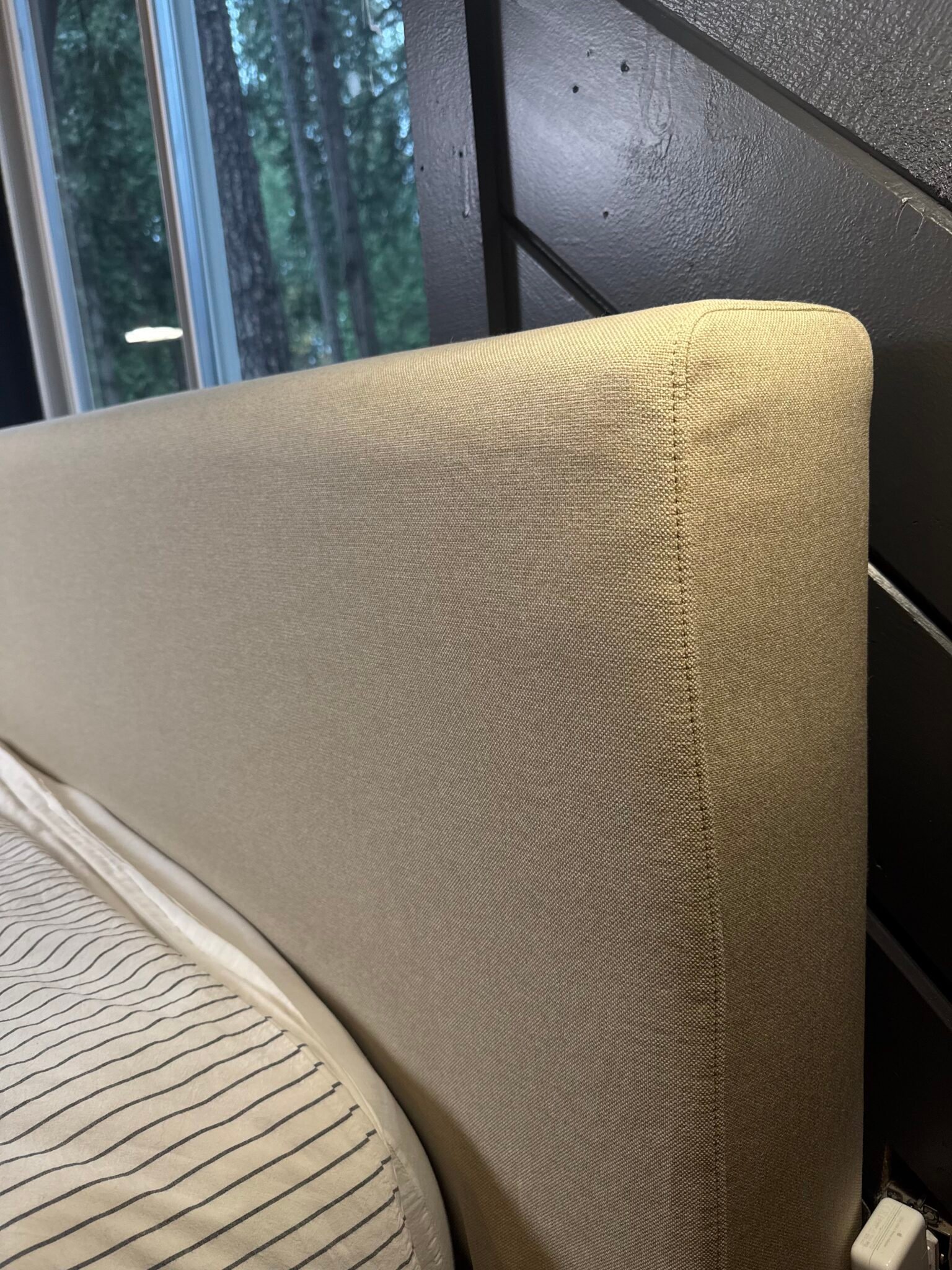 Close-up of a beige upholstered headboard next to a striped mattress, with a window and dark wooden wall in the background.