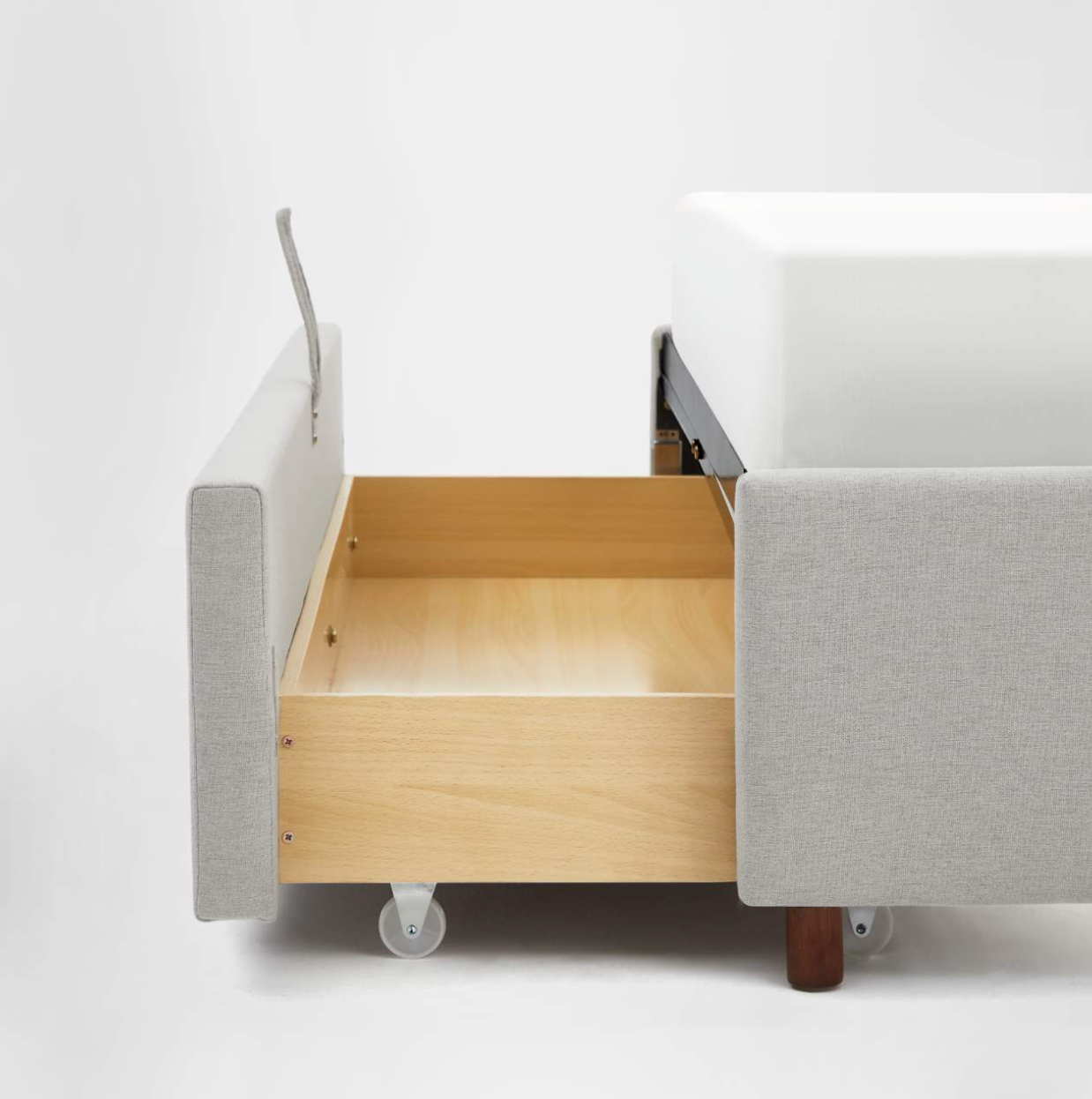 A close-up of a light grey upholstered bed with an open wooden storage drawer on wheels underneath the mattress.