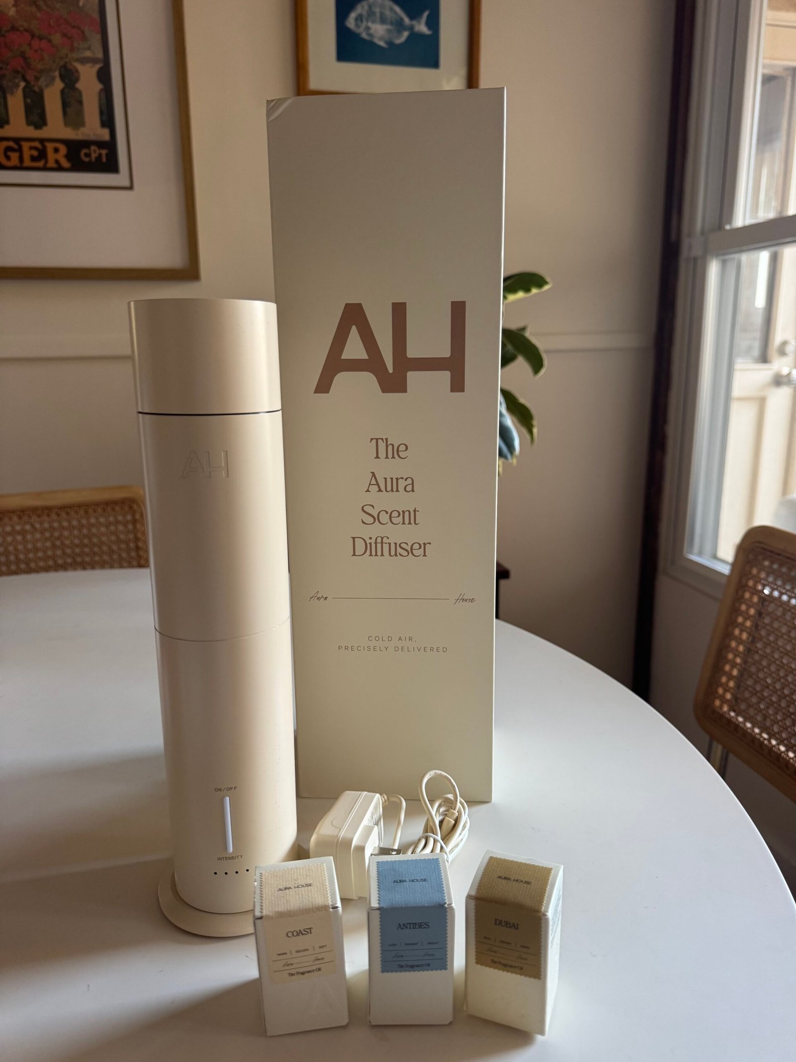 A white scent diffuser with its box is on a table, next to three scent capsules labeled Coast, Artemis, and Balm, in a room with art on the walls and a window.