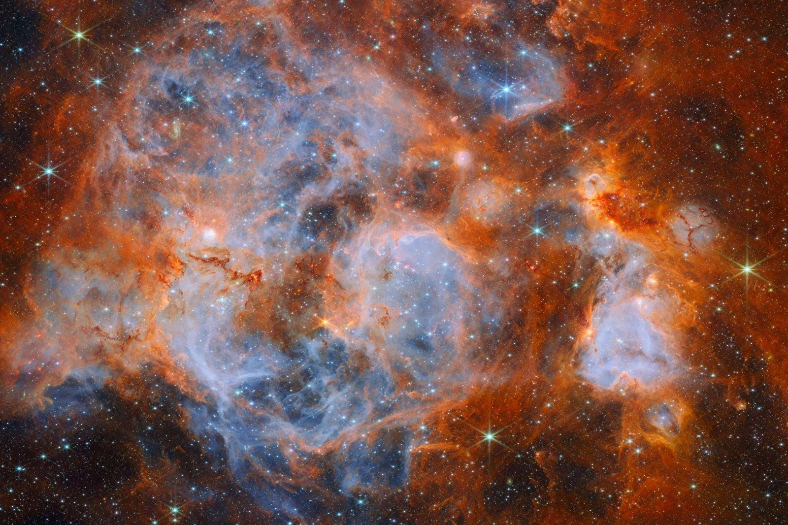 A nebula made of clouds of gas and dust with stars forming within. In the center and out to the left it is filled with thin, wispy blue gas that forms delicate layers. Around this are thicker orange clouds with dark ridges. Thin red filaments of gas cross the center of the nebula. Glowing bubbles in the gas contain dense, very bright clusters of stars. In the edges, distant stars are visible as bright points on a dark background.