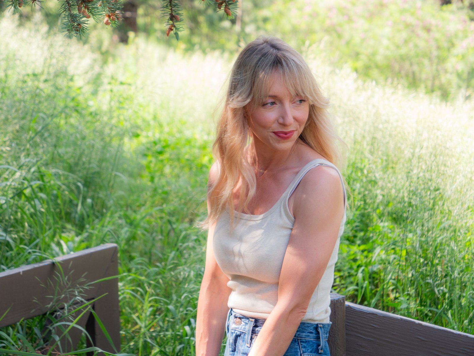 A woman with long blonde hair sits outdoors on a wooden bench, surrounded by tall green grass and trees, wearing a light tank top and jeans.