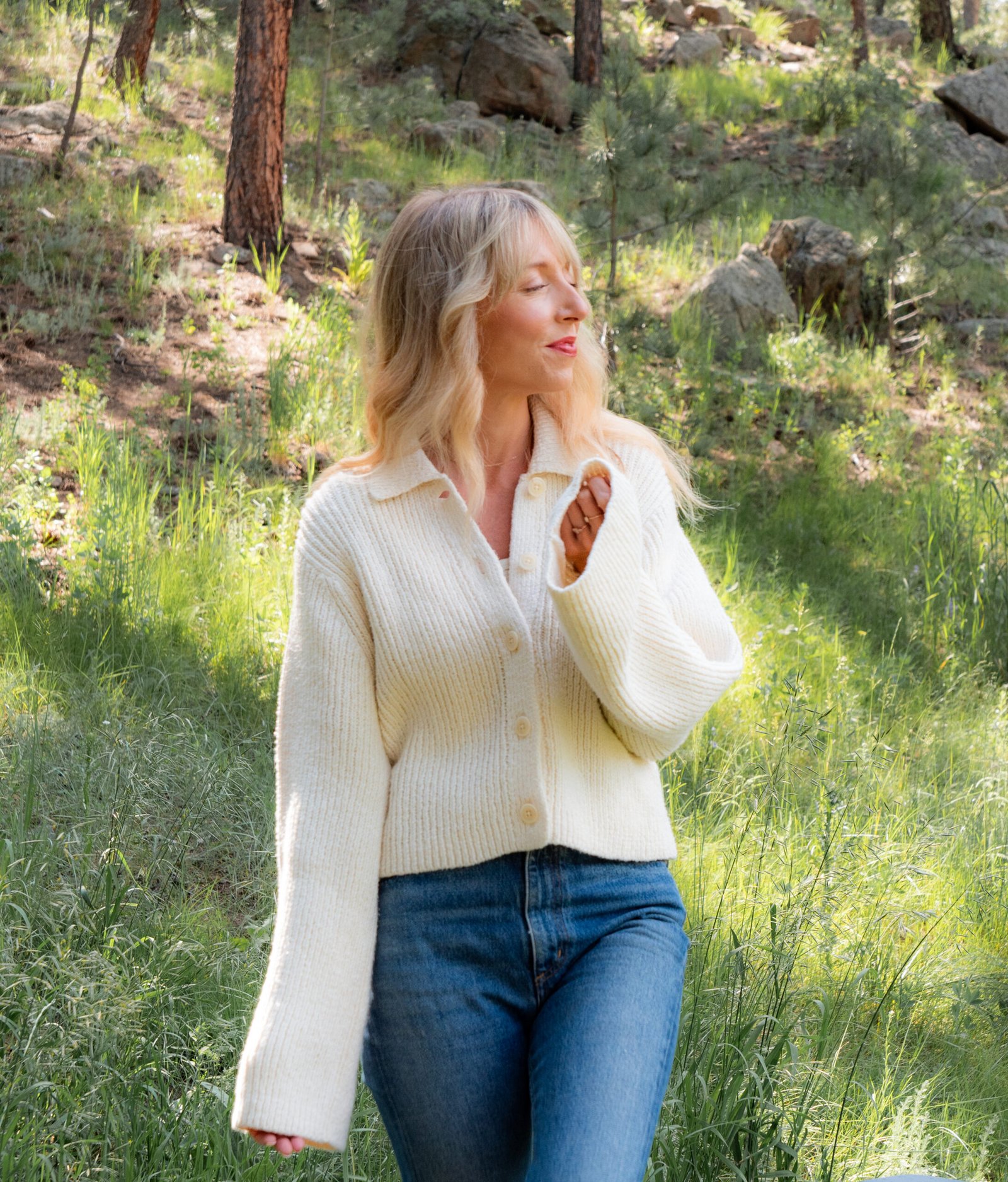 A person with blonde hair wearing a white sweater and blue jeans stands outdoors in a sunlit wooded area with green grass and trees.