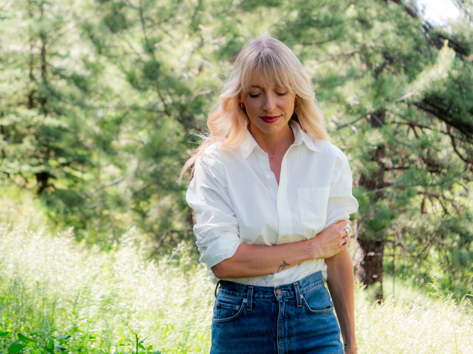 A woman with long blonde hair wearing a white shirt and blue jeans stands outdoors in a grassy area with trees in the background.