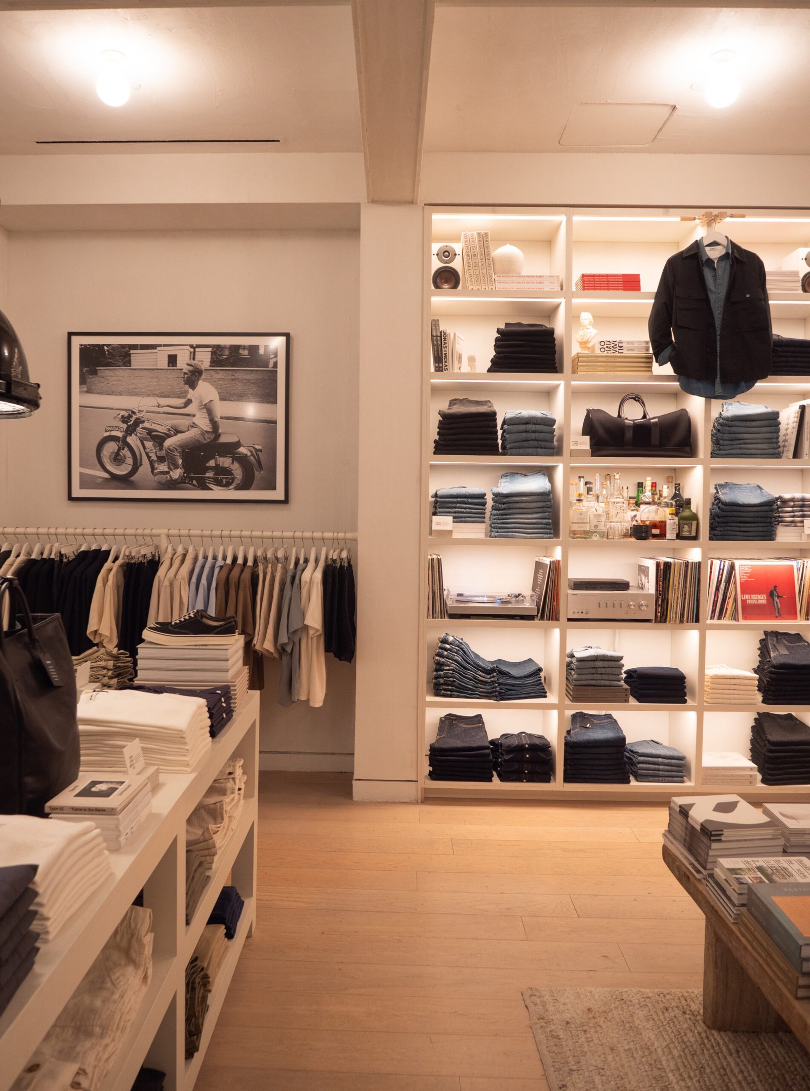 A neatly organized clothing store with folded jeans, shirts, and jackets on shelves, a rack of hanging clothes, and a framed photo of a person on a motorcycle on the wall.