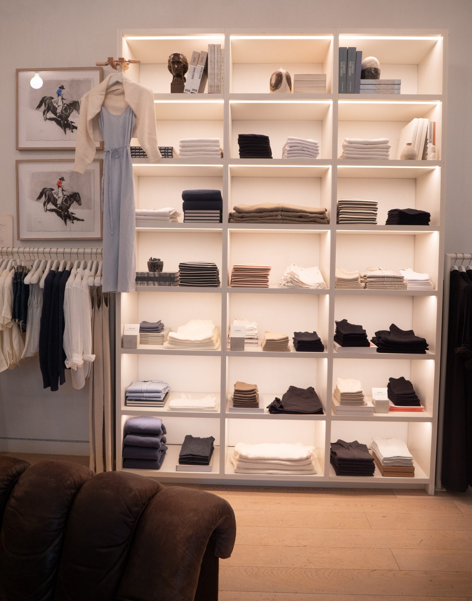 Neatly folded clothes are stacked on illuminated shelves in a retail store, with a dress and scarf displayed on a hanger and framed artwork on the wall.