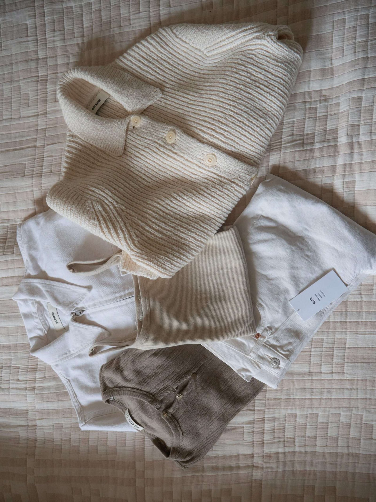 A neatly arranged set of beige and white clothing items, including shirts, pants, and a knit sweater, displayed on a textured quilted surface.