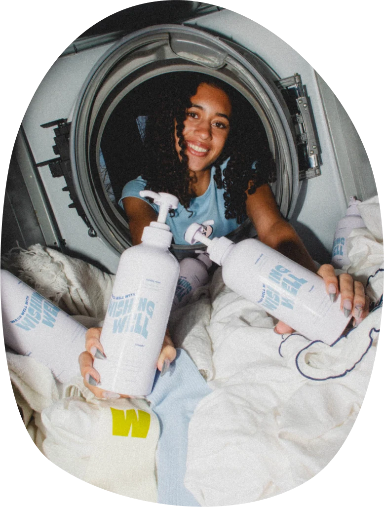 A person sits in a laundry machine surrounded by clothes, holding bottles of laundry detergent and smiling at the camera.
