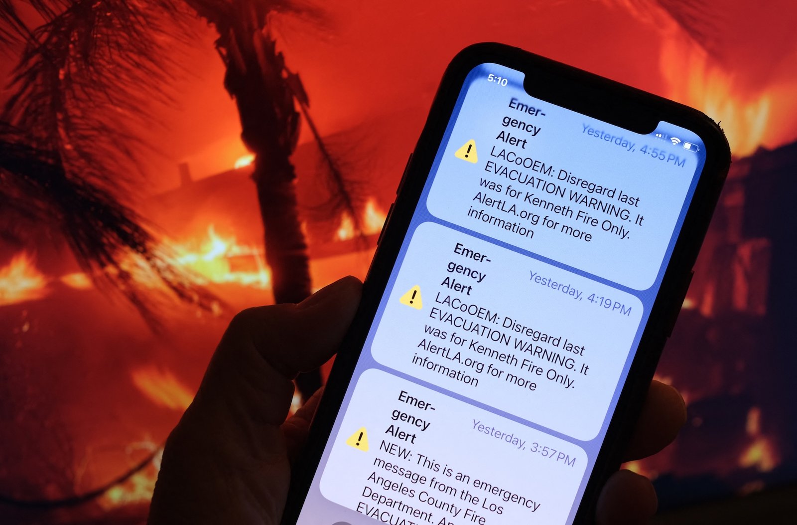 A photo taken January 10, 2025 in Los Angeles, shows the recall message of an evacuation warning issued by the LA County Office of Emergency Management on the screen of a mobile phone in front of a screen showing a photo of the Eaton Fire.